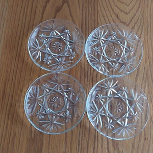 May include: Four clear glass coasters with a starburst design. Each coaster features a central circular pattern surrounded by radiating star-shaped elements. The coasters are arranged on a wooden surface.
