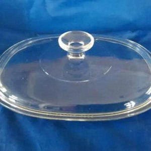 May include: A clear, oval-shaped glass lid featuring a smooth, transparent surface and a centered, rounded glass knob handle. The lid has a slightly raised rim around the edge, designed to fit securely onto a corresponding oval-shaped cookware vessel.