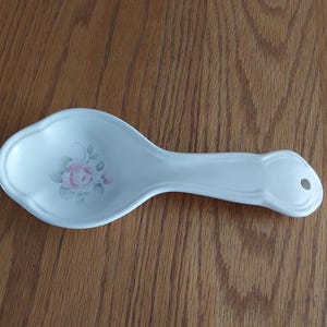 May include: A white ceramic spoon rest with a scalloped edge and a small hole in the handle for hanging. The center of the bowl features a delicate illustration of a single pink rose with green leaves, providing a soft floral accent to the piece.