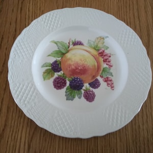 May include: White ceramic plate with a raised woven pattern around the edge. The center of the plate features a painted design of a peach, blackberries, and red currants.