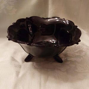 May include: A dark, glossy, black glass bowl with a scalloped rim and three small feet. The bowl has a rounded shape and reflects light, suggesting it is made of a smooth material. The bowl is likely vintage.