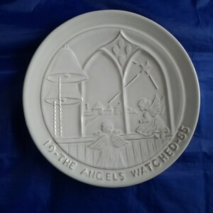 May include: White decorative plate with a relief design featuring angels, a bell, and a church window. The text "19-THE ANGELS WATCHED-85" is inscribed around the edge. The plate is set against a blue background.