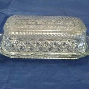May include: A clear glass butter dish with a rectangular shape and a diamond pattern. The dish has a lid and two handles on the sides. The butter dish is sitting on a blue surface.