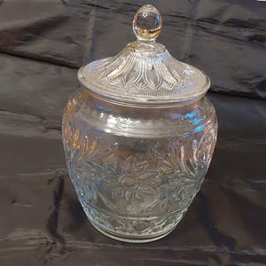 May include: A clear glass jar with a lid, featuring embossed floral patterns. The jar has a round body and a decorative lid with a clear, round knob. The glass has a vintage look, suitable for kitchen or home decor.