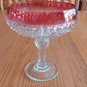 May include: A clear glass footed bowl with a ruby red rim. The bowl has a diamond-cut pattern on the lower portion and a clear stem and base. The bowl is sitting on a wooden table.