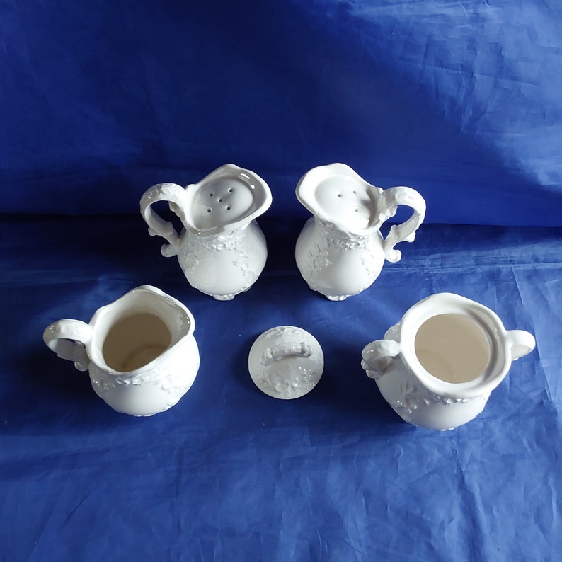 May include: Set of five white ceramic salt and pepper shakers with a floral design. The set includes two shakers with lids, two shakers without lids, and a separate lid.