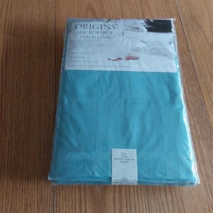 May include: A packaged turquoise microfiber tablecloth. The packaging includes the brand name "ORIGINS" and the product description. The tablecloth is folded and has a textured surface. The packaging is sealed in clear plastic.