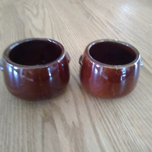May include: Two small brown ceramic bowls with rounded edges and small handles on each side.