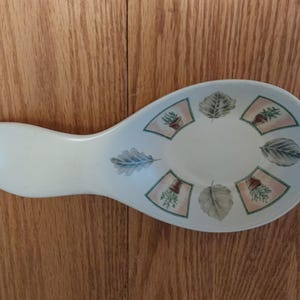 May include: Off-white ceramic spoon rest with a leaf and acorn design. The rest has a wide, oval-shaped bowl with a handle. The design includes green and tan leaves and acorns, with a light pink border.