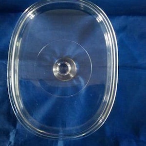 May include: A clear, oval-shaped glass lid with a silver-colored handle in the center. The lid has a rounded edge and is designed to fit a pot or pan. The transparent design allows visibility of the contents.