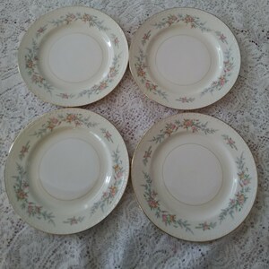May include: Set of four white ceramic dinner plates with a floral design in shades of pink, green, and yellow. The plates have a gold trim around the edge.