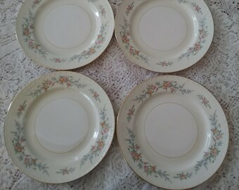 May include: Set of four white ceramic dinner plates with a floral design in shades of pink, green, and yellow. The plates have a gold trim around the edge.
