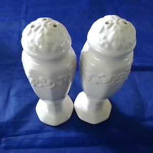 May include: A set of two white ceramic salt and pepper shakers with an ornate design. The shakers have a round top with three holes for dispensing salt and pepper.