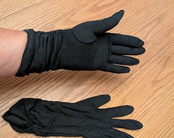 May include: A pair of black gloves made from a soft, stretchy material. One glove is on a person's hand, the other lies flat on a wooden surface. The gloves have a classic design, with a gathered wrist and individual finger slots.