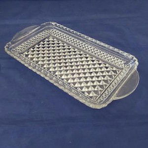 May include: A rectangular clear glass tray with a diamond pattern in the center and a textured border. The tray has two rounded handles on either end. The tray is on a blue surface.