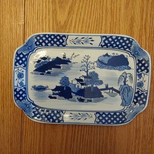 May include: A blue and white ceramic serving tray with a traditional Chinese landscape design. The tray features a blue and white lattice border.