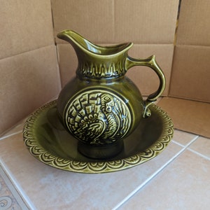 May include: A green ceramic pitcher and bowl set with a raised design of a turkey. The pitcher has a handle and a wide spout. The bowl has a scalloped edge.