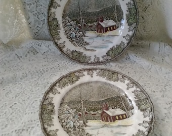 May include: Two vintage ceramic plates with a winter scene of a red schoolhouse in a snowy forest. The plates have a green and brown leafy border.