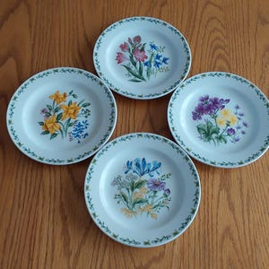 May include: Four white ceramic plates with floral designs. Each plate features a different arrangement of colorful flowers, including yellow, blue, purple, and pink, with a green border. The plates are arranged on a wooden surface.