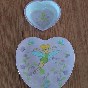 May include: A heart-shaped pink and purple plate with a Tinkerbell illustration. The plate features a green and blue fairy with wings, flying over purple flowers and vines. The text "Pretty little pixie" is written in black on the plate.