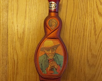 May include: A red glass decanter with a cork stopper. The decanter is shaped like a figure eight and features a label that reads "Beam's Choice". The front of the decanter has a picture of a man wearing a straw hat and a blue shirt.