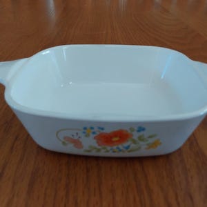 May include: White square baking dish with two handles and floral design. The dish features a pattern of orange, blue, and yellow flowers. A vintage kitchenware item.