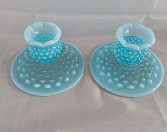 May include: Two light blue glass candle holders. Each has a round base with a textured pattern of small, raised dots. A central, textured, rounded column supports a small, flower-shaped cup. The candle holders are made of glass.