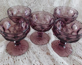 May include: Five vintage amethyst-colored dessert glasses. Each glass features a round bowl with a decorative pattern and a stem. The glasses are arranged on a white lace tablecloth.