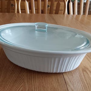 May include: An oval, white ceramic baking dish with a clear glass lid. The dish has a ribbed design on the sides and a light blue rim. The lid has a small, rectangular handle. The dish is on a wooden table.