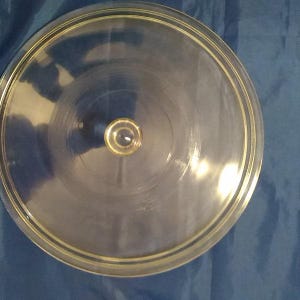 May include: A clear, round glass lid with a gold-colored rim and a small, gold-colored knob in the center. The lid is designed to fit on a pot or pan. The glass is transparent, allowing visibility of the contents.