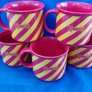 May include: A set of six red coffee mugs with yellow diagonal stripes. Each mug has a handle and the word "Kahlúa" printed on the side in a white, stylized font. The mugs are stacked on a blue surface.