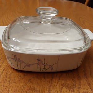 May include: A white square casserole dish with a clear glass lid. The dish has two white handles and a floral design with purple irises. The dish is sitting on a wooden table.