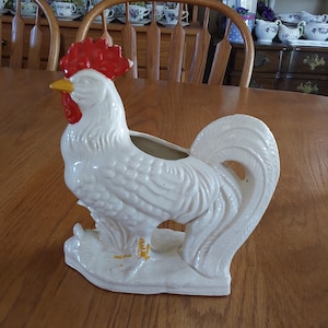 May include: A white ceramic rooster planter with a red comb and wattle, and a yellow beak. The rooster has a detailed feather design and is standing on a base. The planter is a decorative item for home decor.