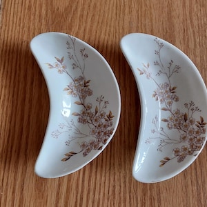 May include: Two off-white, crescent-shaped dishes with a floral design in shades of brown and gold. The dishes are placed on a wooden surface, showcasing their elegant shape and decorative pattern. These dishes are ideal for serving appetizers or small portions.
