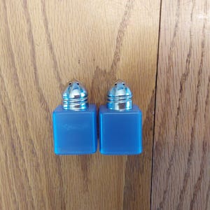 May include: Two blue salt and pepper shakers with silver-colored tops. The shakers are square-shaped and sit on a wooden surface. The tops have a series of small holes for dispensing the contents.