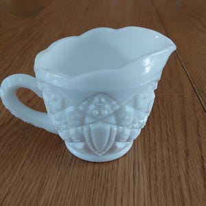 May include: A white, vintage-style creamer with a decorative, embossed pattern. The creamer has a handle and a scalloped rim. The creamer is made of opaque white glass and sits on a wooden surface.
