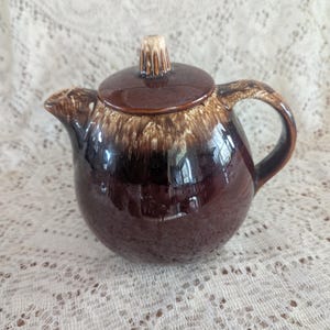 May include: A brown glazed ceramic teapot with a white knob on the lid. The teapot has a brown and white mottled glaze.
