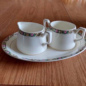 May include: Cream-colored teacup and creamer set with floral and gold trim. The set includes a small creamer pitcher, a teacup, and a matching oval-shaped serving plate. The floral design features pink, green, and yellow flowers.