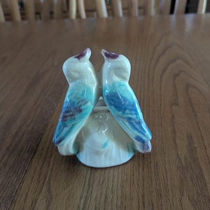 May include: A ceramic salt and pepper shaker set featuring two birds facing each other. The birds have blue and teal wings, with purple accents on their heads. The base is off-white with a cracked texture.