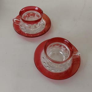 May include: Two clear glass teacups with red rims and matching saucers. The cups and saucers have a decorative pattern. The cups have handles. The saucers are round.