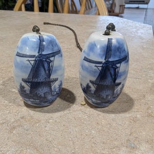 May include: Two blue and white ceramic egg-shaped containers with a windmill design. Each has a metal top and a chain connecting them. The containers are on a light-colored surface.