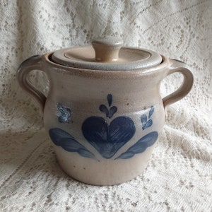 May include: A beige stoneware crock with a lid and two handles. The crock features a blue heart design with leaf accents. The lid has a small knob. This handmade pottery piece is suitable for kitchen storage.
