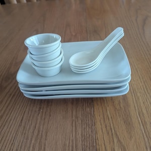 May include: A stack of four white rectangular plates, with a stack of small white bowls and four white soup spoons. The items are arranged on a wooden table. The bowls and spoons are white.