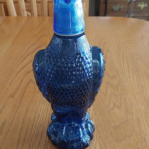 May include: A cobalt blue glass decanter shaped like an eagle. The bottle features a textured surface mimicking feathers across the body and wings, with a removable head piece that serves as a stopper. The glass is translucent and deep blue.
