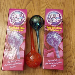 May include: Two pink boxes and two hand-blown glass watering bulbs. The boxes read "Aqua Globes" and "Hand-Blown Glass Watering Bulb." One bulb is red, and the other is green and blue. The boxes have the text "The Work of Art that waters your plants for you!"