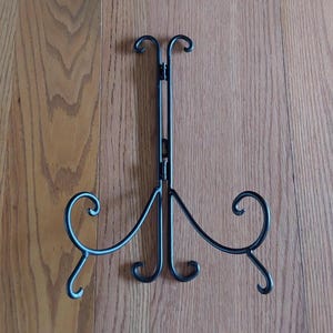 May include: A black metal display stand with a decorative, scrolled design. The stand has three curved legs and a central support that extends upwards, with two curved hooks at the top. The stand is designed to hold a picture or plate.