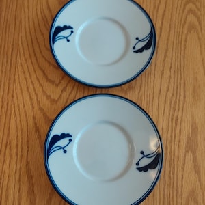May include: Two white ceramic saucers with a blue floral design. The saucers have a raised rim and a small indentation in the center.