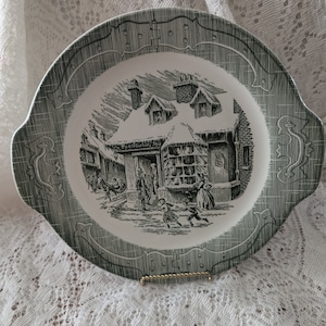 Puede incluir: Un plato de cerámica verde y blanco con una escena de invierno de un edificio con nieve en el techo y personas caminando por la calle. El plato tiene un borde decorativo.