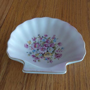 May include: A white ceramic dish shaped like a scallop shell with a scalloped edge and a gold-trimmed rim. The center of the dish features a colorful floral bouquet print with pink, yellow, and blue flowers and green leaves on a white background.