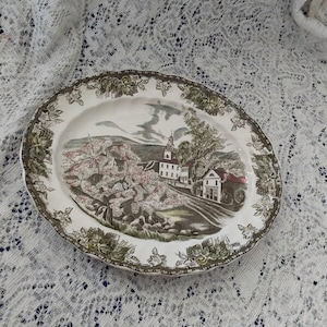 May include: Oval ceramic serving platter with a scenic landscape design. The platter features a central image of a village with a church and flowering trees, framed by a border of green and brown floral patterns. The rim has a scalloped edge.